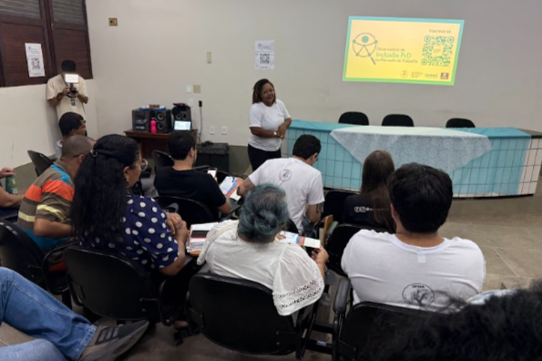 Imagem do auditório anfiteatro da Ufma. É possível ver apenas as costas das pessoas que estão sentadas. Mas é possível ver uma tela de projeção com o logotipo do Observatório, enquanto Sâmia, uma mulher negra de cabelo cacheado e camisa braca, está à frente comentando algo.