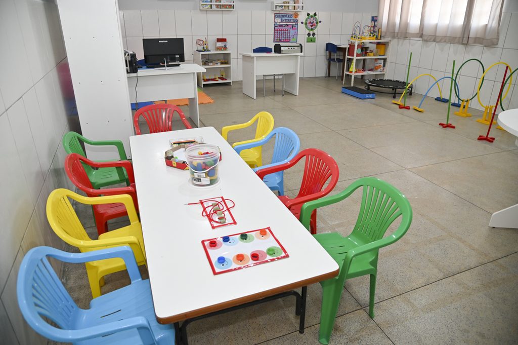 Sala com cadeiras e recursos diversos coloridos. EM primeiro plano. há uma mesa com alguns materiais e 8 cadeiras coloridas. Ao fundo, há recursos guardados em armários. Fim da descrição.